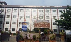 A view of the RNS Vidyaniketan school building, featuring a modern white facade with multiple windows, the school name prominently displayed, a signboard at the entrance, and a well-maintained garden in the foreground.