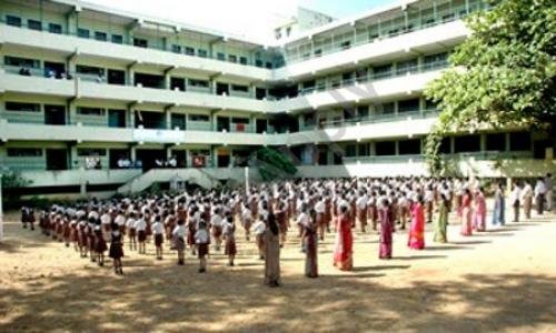 Students of Sri Sarvajna Public School participating in a morning assembly in the open courtyard of a spacious multi-story school building with lush greenery on the side.